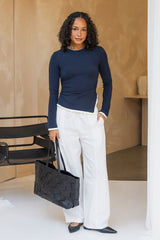A woman stands indoors wearing a fitted navy long-sleeve top, white wide-leg pants, and black pointed flats, while holding a large black woven tote bag. The background features a warm beige textured wall and a modern black chair.