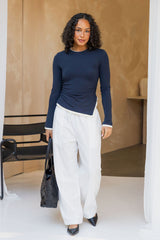 A woman modeling a navy blue long-sleeve top and white wide-leg pants, holding a black handbag in a modern indoor setting with a beige textured wall.