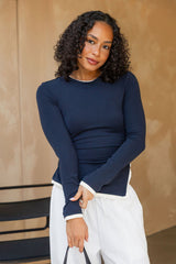 A woman with curly dark hair wearing a navy blue long-sleeve top with white contrast trim and white pants, standing against a warm beige textured wall.