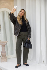 Woman taking a selfie in front of a mirror wearing a black leather jacket and green pants.