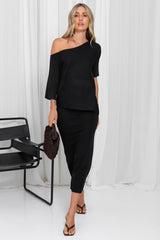 A woman models a black two-piece outfit consisting of an off-the-shoulder short-sleeve top and a matching midi skirt, holding a brown textured clutch bag against a white curtain backdrop.