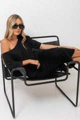 A woman wearing a black one-shoulder dress and black sunglasses sits in a black metal lounge chair against a plain white studio background.