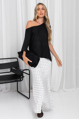 A woman models a black off-the-shoulder knit top paired with a white maxi skirt featuring a black polka-dot pattern, holding a black clutch bag in a studio setting with white drapes.