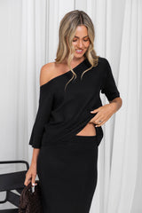 A woman modeling a black one-shoulder long-sleeve dress with a twisted waist detail and a side cutout, standing against a white curtain background.