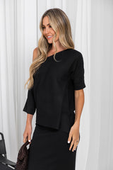 A woman modeling a black off-the-shoulder short-sleeve top with a matching black skirt, standing against a white curtain backdrop.