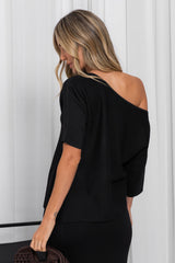 A woman shown from the back wearing a black off-the-shoulder short-sleeve top and black bottoms, holding a dark brown woven handbag against a white paneled background.