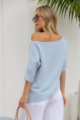 A woman wearing a light blue off-the-shoulder knit top and white pants, photographed from the back in a bright indoor setting with a white wall and green leaves at the top.