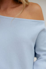 Close-up of a person wearing a light blue off-the-shoulder ribbed top, showing the shoulder, collarbone, and part of the sleeve against a plain light background.
