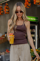 A woman wearing a dark brown ribbed tank top and beige plaid shorts stands in a grocery store holding a cup of orange juice. She is wearing sunglasses and a delicate necklace.