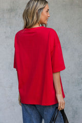 A woman shown from the back wearing an oversized bright red short-sleeve t-shirt and blue jeans, holding a black bag against a textured gray concrete wall.