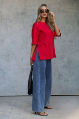 A woman modeling a bright red short-sleeve top with side slits, paired with wide-leg blue jeans, black sunglasses, and a black handbag, standing against a textured gray concrete wall.