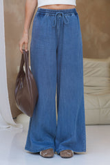 A person wearing wide-leg blue denim pants with an elastic drawstring waist, holding a brown leather handbag, standing in an indoor setting with a beige sofa in the background.