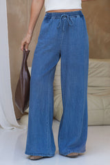 A person wearing wide-leg blue denim drawstring pants, holding a brown leather bag, standing in a brightly lit indoor setting with a beige sofa in the background.