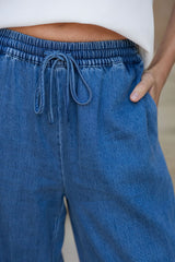 Close-up of a person wearing blue denim drawstring pants with a white top, showing the waistband, drawstring, side pocket, and hand.