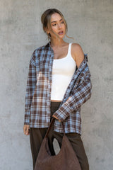 Woman wearing a plaid shirt over a white tank top, holding a brown bag against a concrete wall.