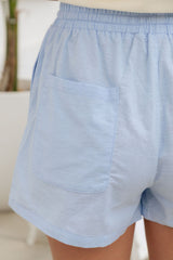Close-up of a person wearing light blue linen-blend shorts with an elastic waistband and a large patch pocket on the back.