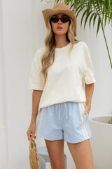 A woman modeling a casual outfit featuring an oversized cream-colored short-sleeve t-shirt and light blue shorts with white side stripes, accessorized with a straw hat, sunglasses, and a woven bag.
