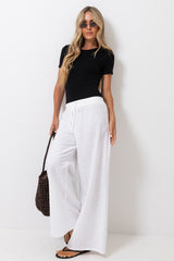 A woman modeling white wide-leg drawstring pants with a black short-sleeve top, sunglasses, and a brown woven handbag against a plain white studio background.