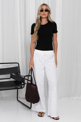 A woman stands in a studio wearing a fitted black short-sleeve top, white wide-leg drawstring pants, sunglasses, and sandals, while holding a dark woven tote bag. A black chair is visible at left against a white draped backdrop.
