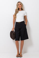 A woman stands against a plain white wall wearing a fitted white short-sleeve top, black knee-length pleated shorts with a thin black belt, and black sandals. She holds a dark woven handbag.