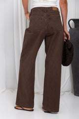 A person shown from the back wearing dark brown wide-leg jeans, a white top, and brown sandals, holding a black woven bag against a white curtain backdrop.