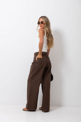 A woman wearing brown wide-leg pants, a white tank top, and sunglasses stands in profile against a plain white studio background.