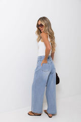 A woman modeling a pair of light-wash wide-leg denim jeans, paired with a white tank top and sunglasses, standing against a plain white studio background.
