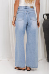 A person shown from the back wearing light-wash wide-leg denim jeans, a white top, and brown sandals in a studio setting with a white curtain backdrop and a dark vase on the right.