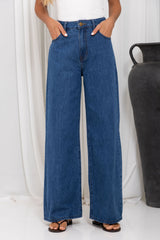 A person wearing high-waisted, wide-leg blue denim jeans with a white top and brown sandals, standing in front of a white curtain with a large dark vase on the right.
