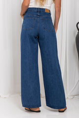 A person shown from the back wearing high-waisted, wide-leg blue denim jeans with a white top and black sandals, standing against a white draped studio backdrop.