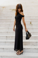 A woman wearing a black off-the-shoulder maxi dress stands on white stone steps, holding a black handbag and wearing black strappy sandals.