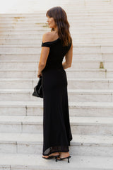 A woman stands on outdoor stone steps wearing a black off-the-shoulder maxi dress, black pointed-toe heels, and holding a black handbag.
