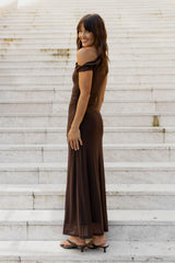 A woman wearing a dark brown off-the-shoulder maxi dress and black heeled sandals, standing on light-colored stone steps and looking back over her shoulder.