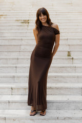 A woman wearing a dark brown off-the-shoulder maxi dress stands on light-colored stone steps.