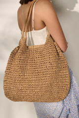 A large woven straw tote bag with double shoulder straps, carried over the shoulder of a model wearing a white camisole and a blue floral skirt against a light wall with soft shadows.