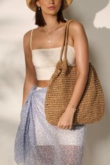 A woman wearing a straw hat, white crop top, and a blue floral sarong carries a large woven straw tote bag against a sunlit neutral wall.