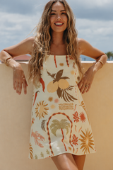 A woman wearing a sleeveless mini dress with a tropical print, leaning against a beige wall outdoors under a cloudy sky.