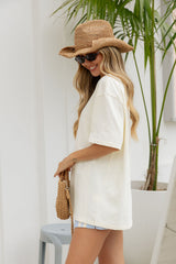 A woman wearing an oversized off-white short-sleeve T-shirt, a straw hat, sunglasses, and a woven handbag, standing in a bright indoor setting with a potted palm plant.