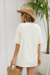 A woman viewed from behind wearing an oversized off-white t-shirt, a straw hat, and holding a woven straw bag, standing next to a potted palm plant.
