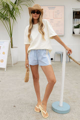 A woman models a casual summer outfit featuring an oversized cream t-shirt, light blue shorts with white side stripes, a straw hat, and beige sandals, standing in a bright, modern outdoor setting.