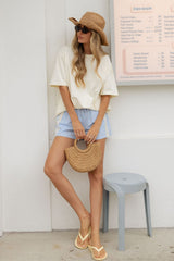 A woman stands outdoors wearing a cream oversized T-shirt, light blue denim shorts, a straw hat, sunglasses, and yellow flip-flops, holding a round woven straw handbag. A gray stool and a shop menu board are visible in the background.