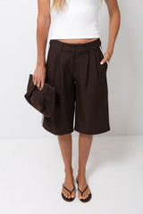 A model wearing dark brown pleated culotte shorts with a button waistband, styled with a white crop top, brown sandals, and holding a matching dark brown clutch bag against a white studio background.