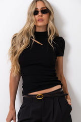 A woman with long blonde hair wearing a black short-sleeve mock neck top with a diagonal cutout at the chest, styled with black trousers and a black belt with a gold buckle, posing against a white wall.