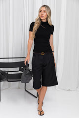 A woman modeling a black short-sleeve mock neck top, black pleated knee-length shorts with a thin black belt, and black strappy heels, standing in front of white draped curtains.