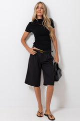 A woman modeling a black short-sleeve mock neck top, black wide-leg cropped shorts, a black belt with a gold buckle, and black flip-flops, holding a black handbag against a plain white studio background.
