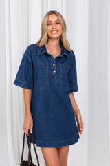 A woman modeling a short-sleeve blue denim dress with a buttoned chest and two front pockets, standing against a white curtain backdrop.