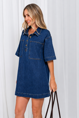 A woman wearing a dark blue denim short-sleeve shirt dress, holding a dark brown braided handbag against a white curtain background.
