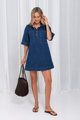 A woman modeling a short-sleeve blue denim dress with a buttoned chest pocket, standing in front of a white curtain backdrop and holding a brown woven tote bag.
