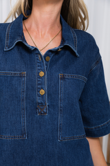 Close-up of a person wearing a dark blue denim short-sleeve collared shirt dress with brass buttons and chest pockets.