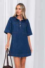 A woman modeling an oversized dark blue denim shirt dress with short sleeves, a buttoned collar, and front patch pockets, standing against a white curtain backdrop and holding a dark woven handbag.